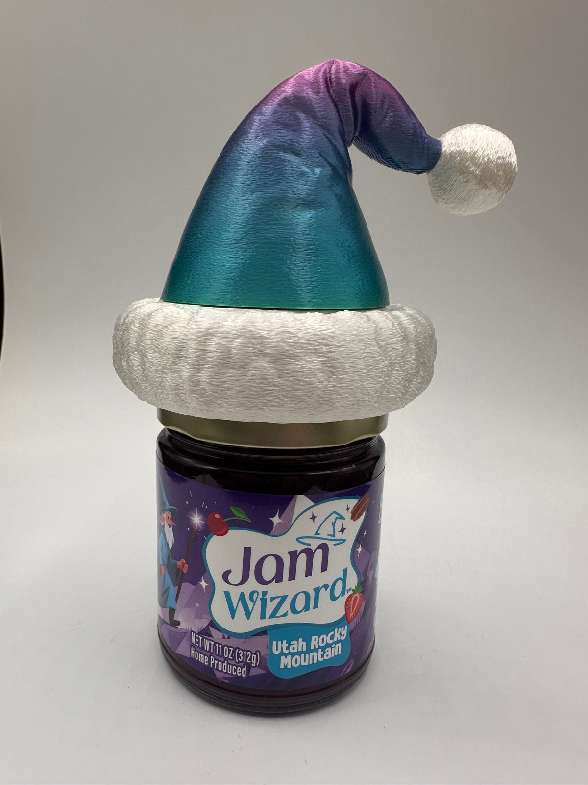 A jar of Jam Wizard Rocky Mountain Jam with a festive Santa hat decoration in the color of Mountain Mirage on the lid, photographed against a plain light gray background.